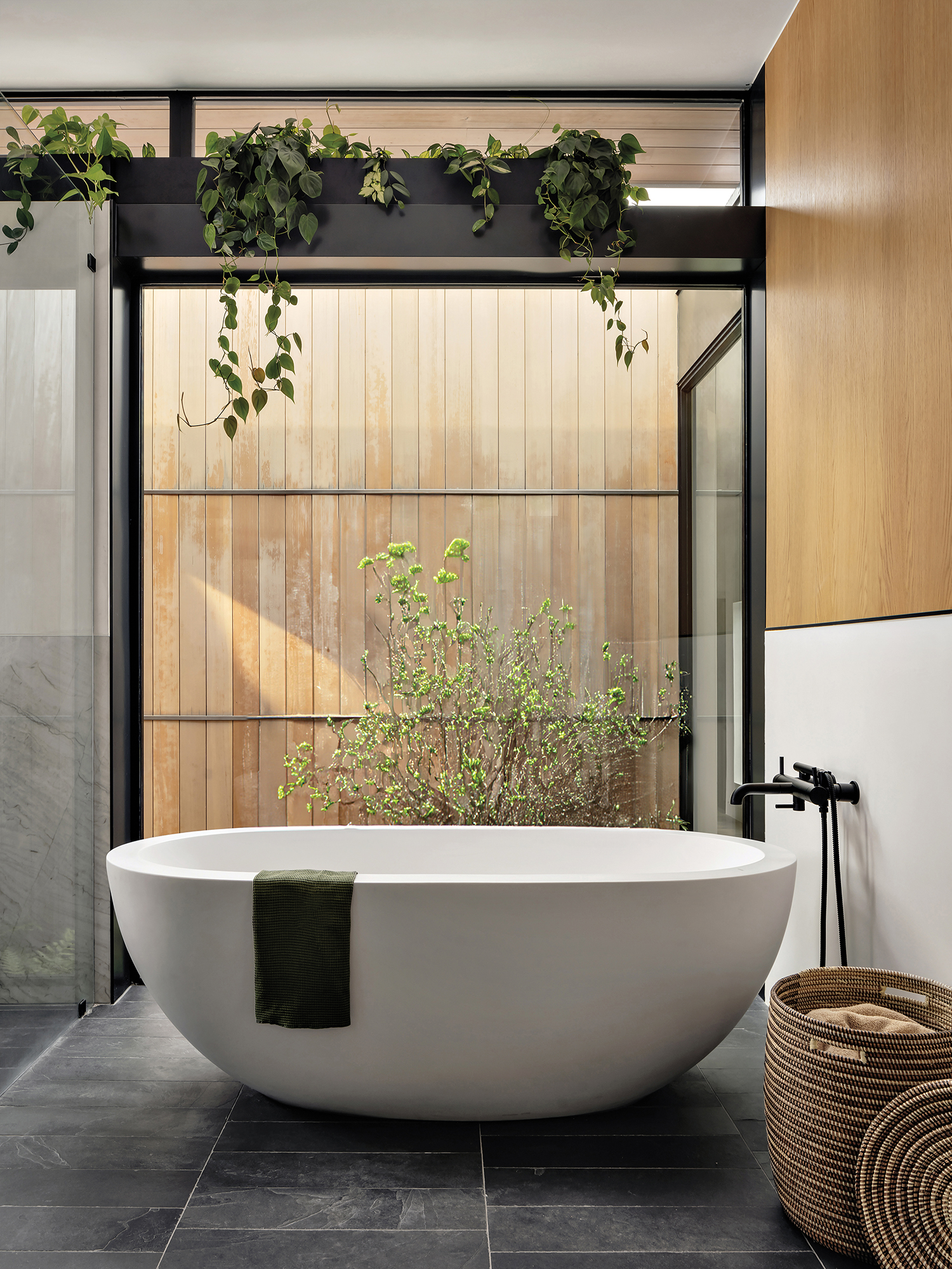 modern bathroom with gray tile flooring, a tub next to a glass wall and plants dispersed throughout