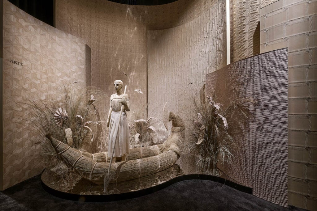 A mannequin in a white dress stands in a woven boat surrounded by dry grasses and textured beige panels in an artfully designed display.