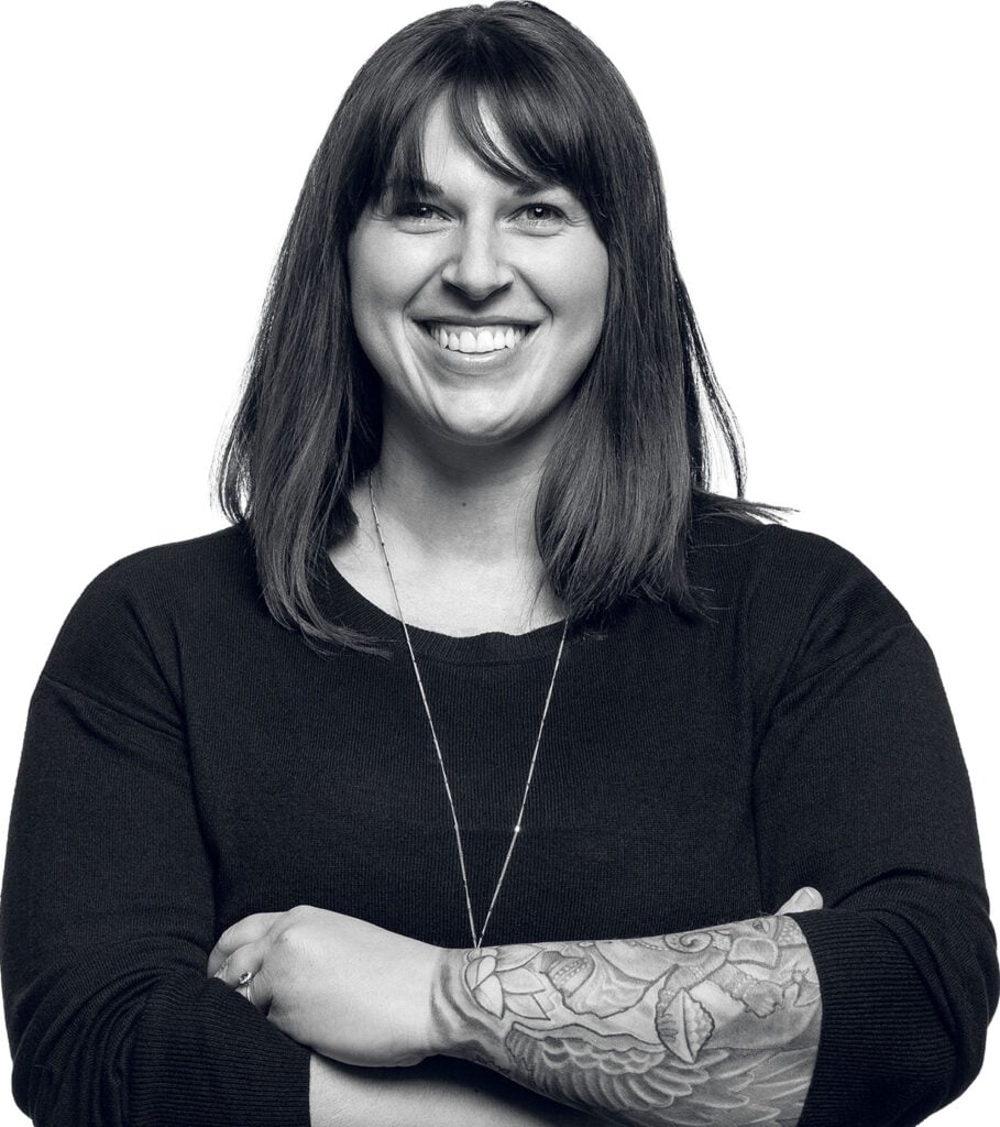 A woman with long dark hair and a tattooed forearm stands smiling with her arms crossed, wearing a dark long-sleeved shirt and a necklace.